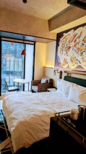 Contemporary hotel room with a large bed, colorful artwork, and a sitting area near the window.