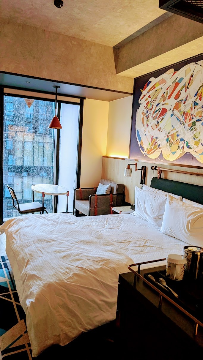 Contemporary hotel room with a large bed, colorful artwork, and a sitting area near the window.
