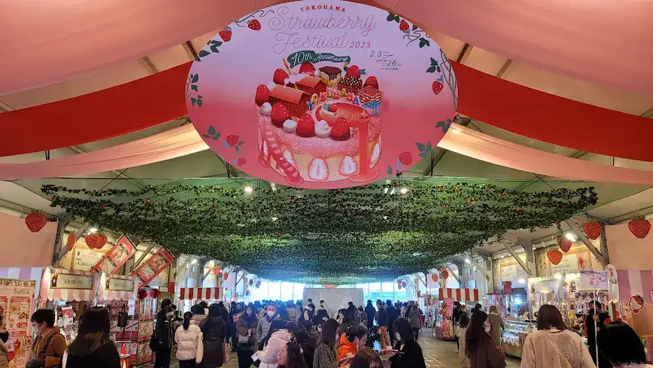 Crowded indoor Yokohama Strawberry Festival 2023 tent with red-and-pink ribbons, strawberry decor, and food stalls.