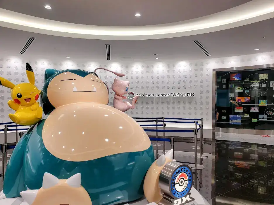 Large Snorlax, Pikachu, and Mew statues at Pokémon Center Tokyo. A person walks past colorful wall displays. Bright and playful atmosphere.