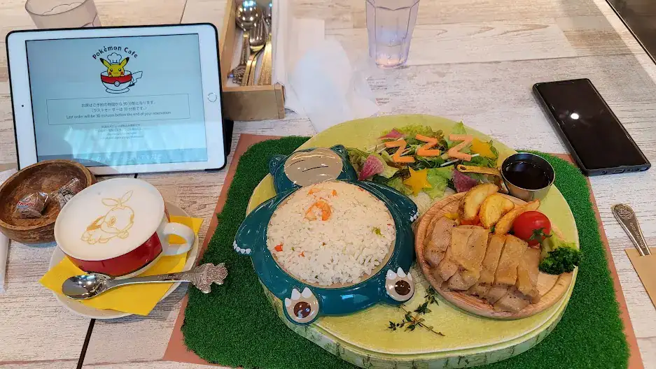 A tablet shows "Pokémon Cafe." A Snorlax dish with rice and a turtwig latte with a Pokémon design sit on a table with utensils nearby.