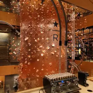 Copper barrel with floral patterns in a coffee shop. Espresso machine with cups in foreground. Warm lighting and modern decor. Text: "R".