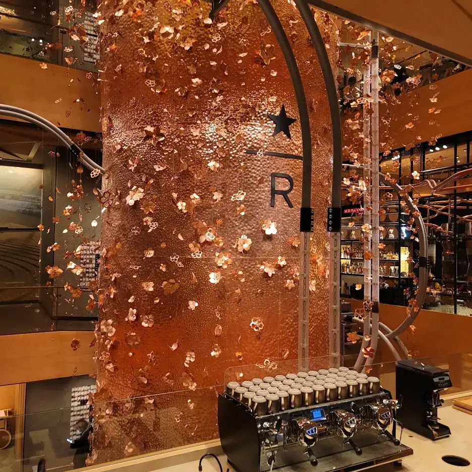 Copper barrel with floral patterns in a coffee shop. Espresso machine with cups in foreground. Warm lighting and modern decor. Text: "R".