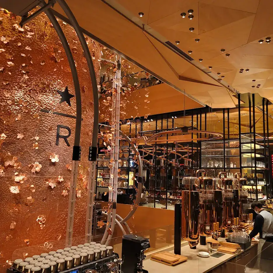 Copper-toned interior with floral patterns on a large wall. A coffee machine on a counter, cups on top. Star and "R" logo visible.