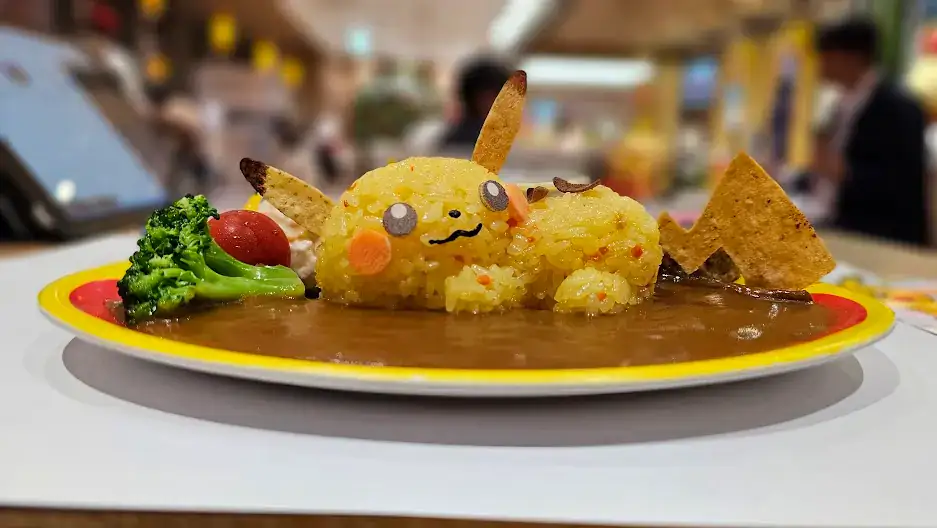 Yellow Pikachu-shaped rice with ears made of chips on curry sauce, surrounded by broccoli and a cherry tomato on a colorful plate.