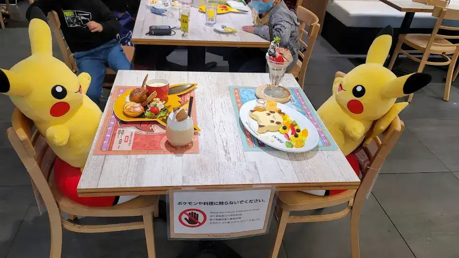 Plush Pikachu toys on chairs at a cafe table. They have a full meal on display. Bright colors and playful decor fill the background.