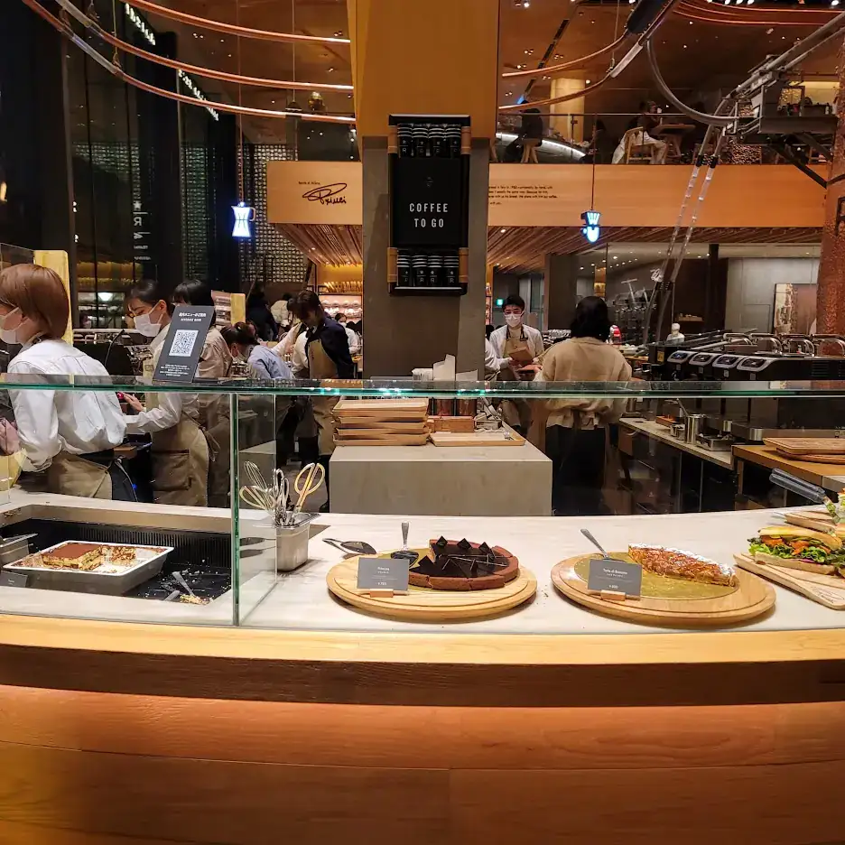 Bakery with staff in masks behind a counter displaying cakes and sandwiches. "Coffee To Go" text above. Warm lighting and busy atmosphere.