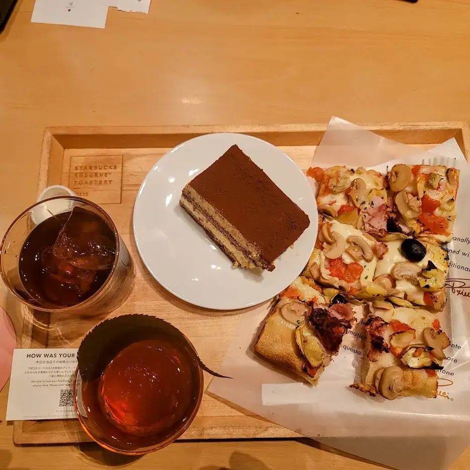 A tray with iced coffees, a slice of tiramisu, and pizza squares on a wooden table. Text on a card reads "HOW WAS YOUR." Warm, cozy setting.