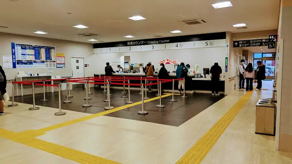 People at a ticketing counter in a brightly lit station with red queue barriers. Signs and schedules are visible on the walls. Quiet atmosphere.