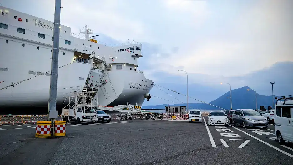 Large white ferry "Sunflower" docked at port with parked cars nearby. Mountain in background under cloudy sky, creating a serene mood.