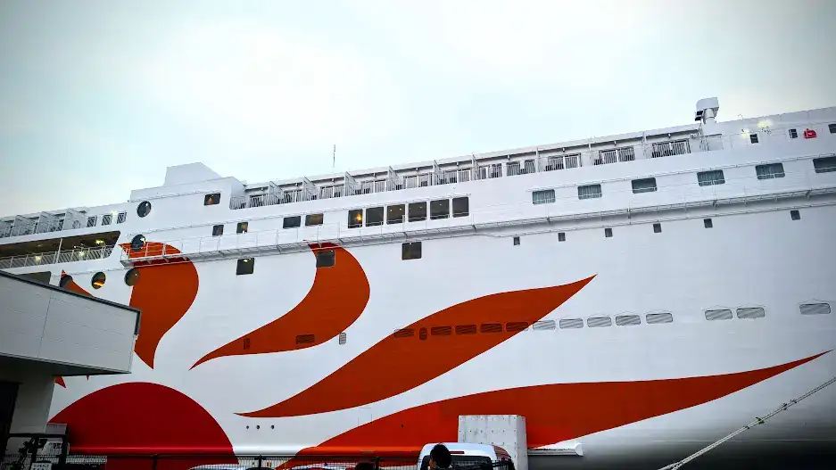 Large white ship with orange and red sun design docked at a port. Pale blue sky in the background. A few structures and vehicles nearby.