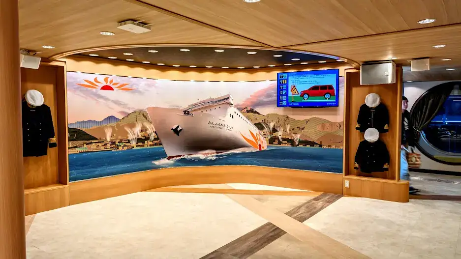 Ship interior with a large mural of a cruise ship at sea. Uniforms are displayed on either side. A screen shows a red car ad. Calm ambiance.
