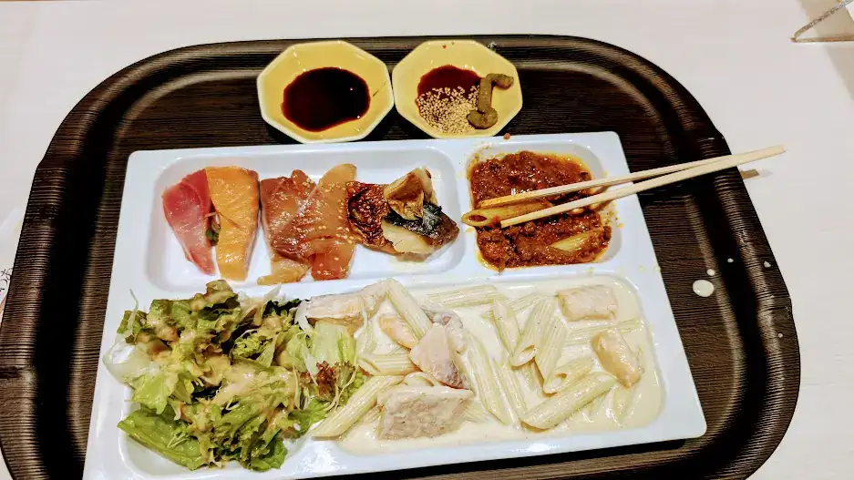 A food tray with salad, assorted sashimi, creamy pasta, and curry. Two bowls of sauces and wasabi on the side, on a dark tray.