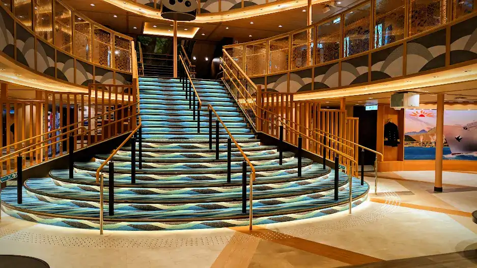 Elegant stairway with wavy blue-patterned carpet in a modern interior, surrounded by golden railings and warm lights. No people present.