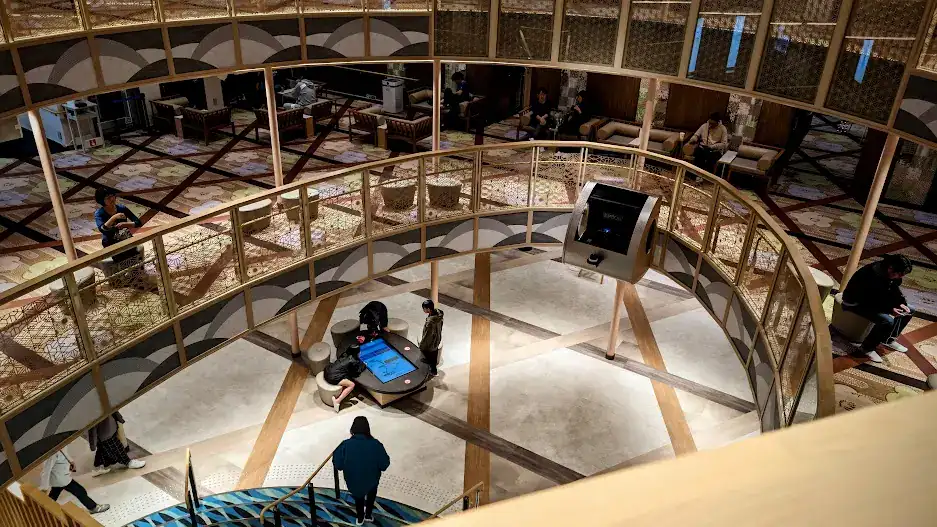 People sit and stand in a modern, circular lounge with patterned floors and gold railings. A young boy interacts with a digital kiosk. View of three decks from above.