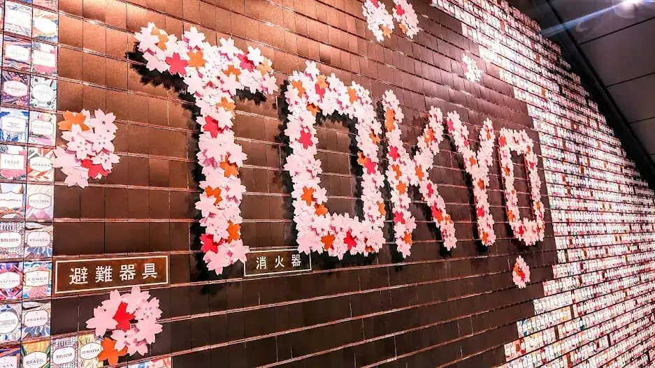 TOKYO spelled out with pink and red flowers on a dark tiled wall. The background features colorful tiles. Text in Japanese is visible.