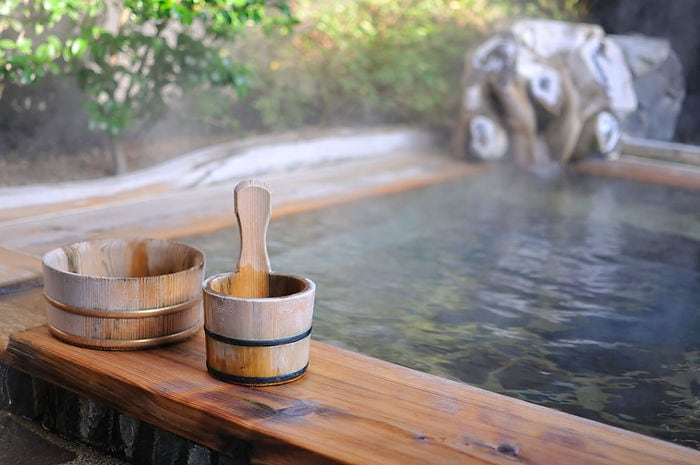 Wooden buckets by a steaming hot spring with trees in the background. The setting is calm and serene, featuring earthy tones.