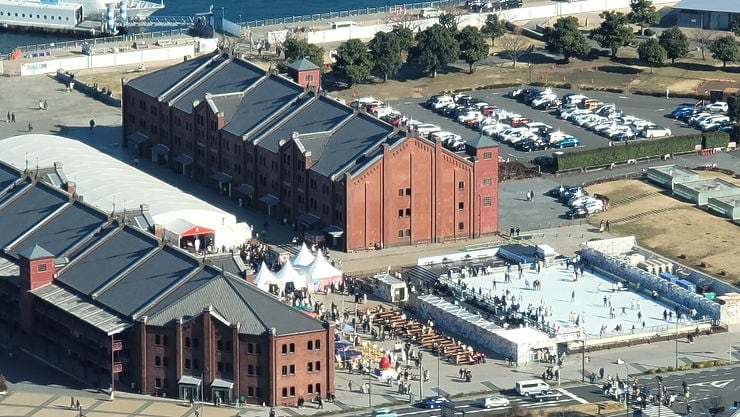 Aerial view of a brick waterfront complex with an outdoor ice rink, crowds skating, and parked cars under a bright blue sky.
