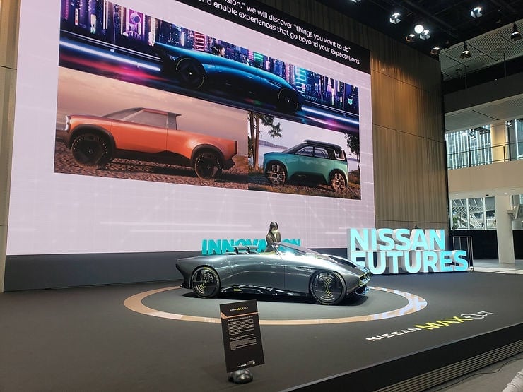 Nissan concept car on stage at a Futures display, with MAXOUT sign, giant screen showing futuristic car designs, and bold white text 