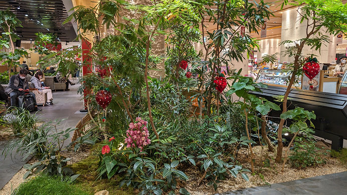 Indoor mall garden with lush plants and red strawberry ornaments; masked shoppers sit nearby, with cafe counters and Japanese signs.