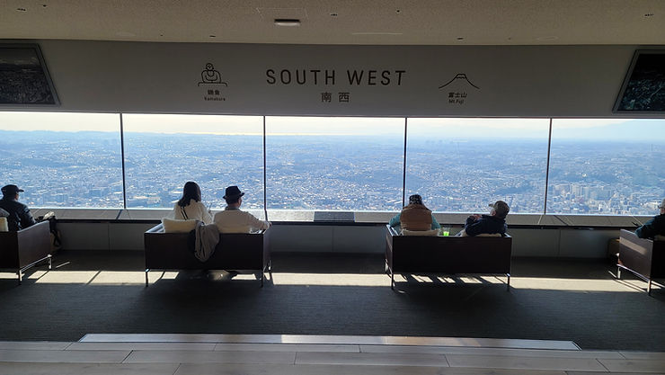 Visitors sit in a lookout lounge under SOUTH WEST, Kamakura and Mt.Fuji signs, gazing over a sunlit city panorama.