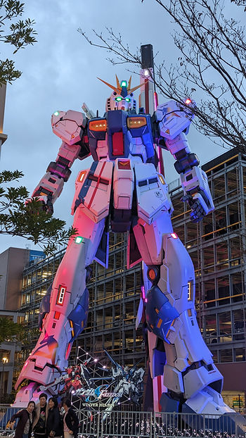 Giant colorful robot statue with glowing lights towers over smiling people. Urban background with buildings and trees. "FREEDOM" text below. RX-93ffν Gundam Statue

