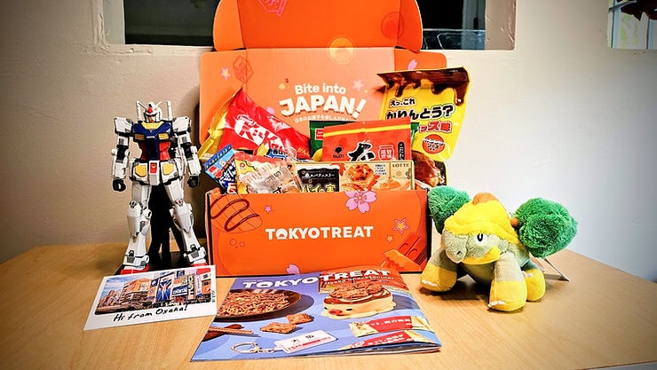 TokyoTreat snack box with Japanese sweets and magazines, flanked by a Gundam figure and green plush toy on a table.