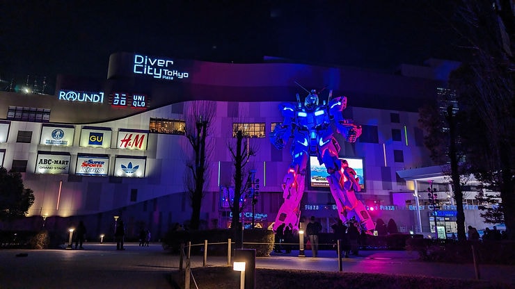 Giant illuminated Gundam statue stands before Diver City Tokyo Plaza at night, surrounded by trees and glowing store signs, creating a vibrant scene.