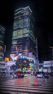 Night scene of Shibuya crossing, Tokyo, with illuminated skyscraper, taxis, and people in vibrant city lights. Rain-wet streets reflect colors.