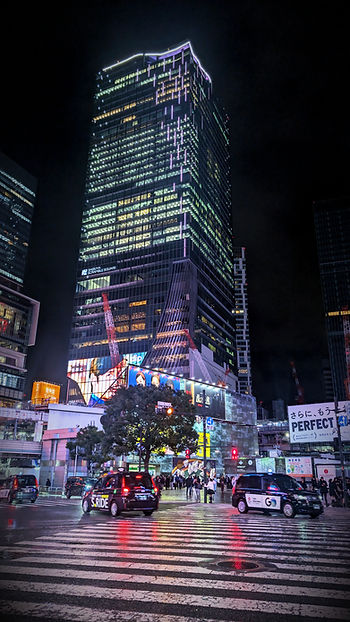 Night scene of Shibuya crossing, Tokyo, with illuminated skyscraper, taxis, and people in vibrant city lights. Rain-wet streets reflect colors.