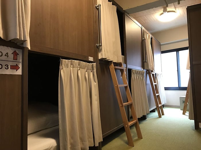 Capsule hotel room with wooden bunk beds and beige curtains. Ladders lead to upper bunks. Room number signs visible. Calm lighting.