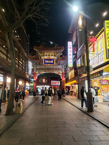 Night street in Chinatown with a brightly lit ornate gate, neon signs, and masked pedestrians walking.