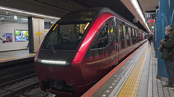 Red train at an indoor station, people on the platform, blue columns, and colorful signs. Mood is calm; no visible text. Hinotori
