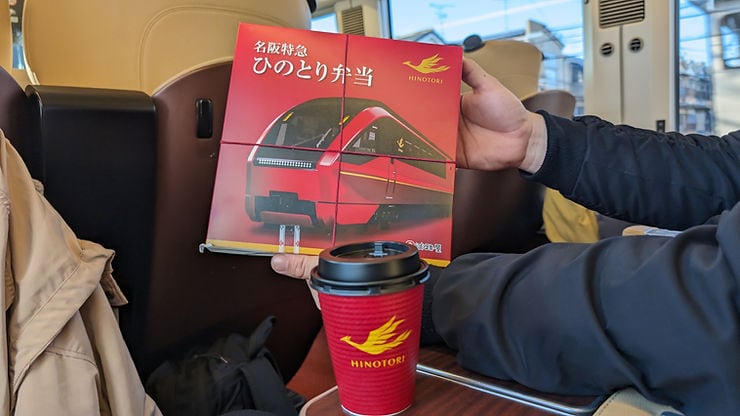 Two hands hold a red Hinotori train-themed box on a train seat, with a red Hinotori coffee cup nearby. Beige and black jackets are visible.