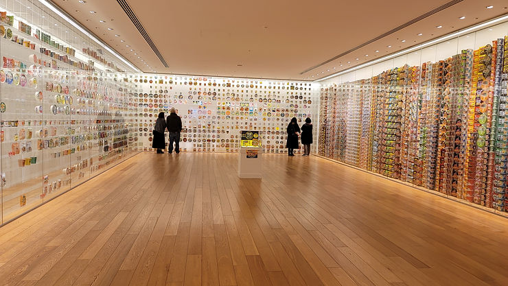 Visitors view walls packed with colorful instant noodle cups in a bright museum gallery with wood floors.
