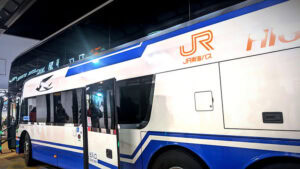 White double-decker bus with blue accents at night, parked at a station. Orange "JR" logo and text visible. Reflective surface shows passengers inside.