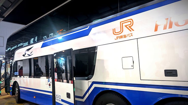 White double-decker bus with blue accents at night, parked at a station. Orange "JR" logo and text visible. Reflective surface shows passengers inside.