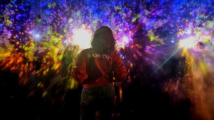 A person in a red jacket stands in front of colorful, abstract lights. The word "HONDA" is visible on their back, creating a vibrant mood.