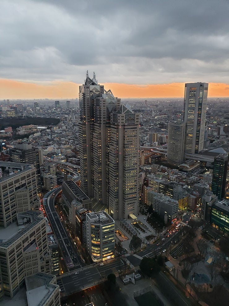 Cityscape with tall skyscrapers under a cloudy sky at dusk. Streetlights and buildings are illuminated, with a vibrant orange horizon.