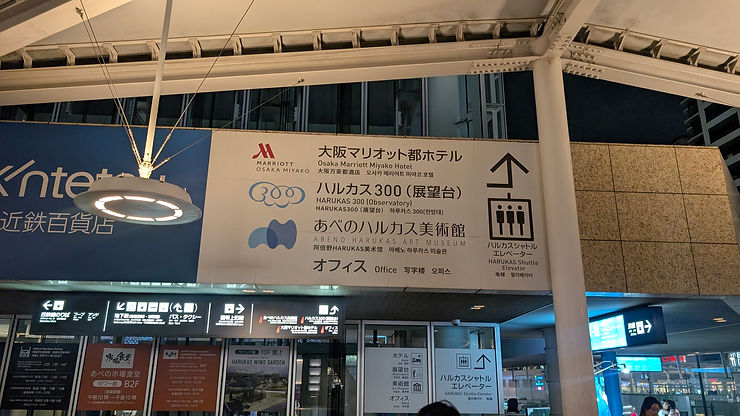 Nighttime in an urban setting with a lit sign for Osaka Marriott Miyako Hotel, Harukas 300, and Abeno Harukas Art Museum. Arrows direct visitors.