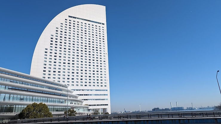 A modern, arched skyscraper hotel under a clear blue sky, with a foreground of a road and greenery. Distant landscape visible on the horizon.