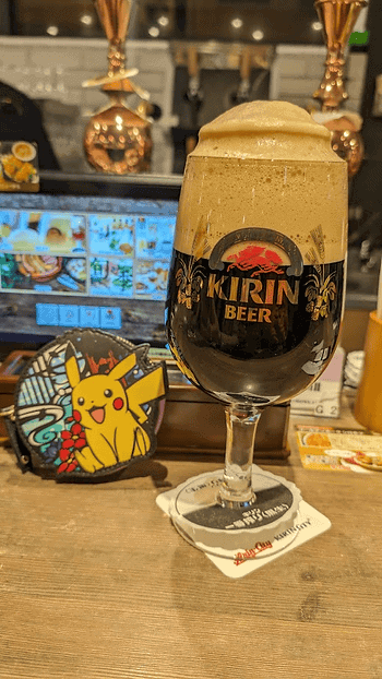 Foamy Kirin beer in a stemmed glass on a bar counter, with a colorful Pikachu sign and menu screens in the background.