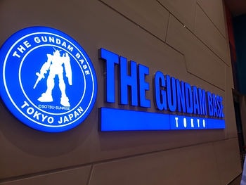 Blue illuminated sign of "The Gundam Base Tokyo" with a Gundam silhouette on a modern wall, conveying a high-tech, vibrant atmosphere.