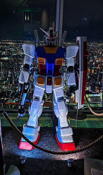 A tall Gundam figure with blue, red, and white colors stands in front of a cityscape at night, illuminated by bright city lights.