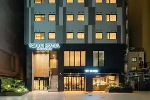 Modern hotel at night with lit windows. "Tosei Hotel Cocone" sign glows neon blue. Plants and soft lighting create a welcoming entrance.