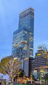 Tall glass skyscraper at dusk with lights on, surrounded by trees decorated with white lights. Signs visible on nearby buildings. Calm ambiance.