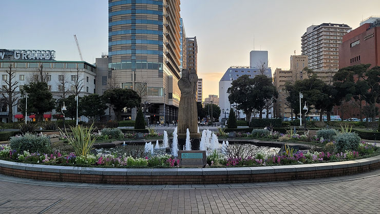City plaza at dusk with a fountain and stone statue in a flowered park, framed by tall buildings and Japanese signs.