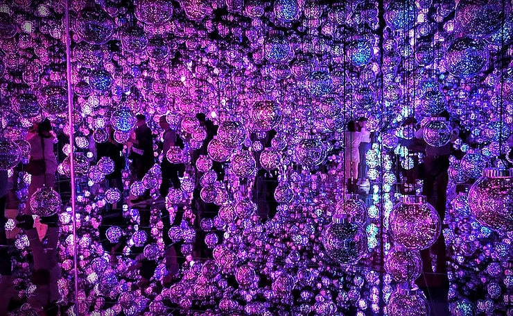 Glowing purple and blue orbs fill a mirrored room, reflecting light. People walk through the space, creating a dreamy, immersive atmosphere.