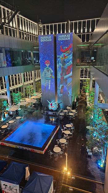 Night scene of a rooftop plaza with colorful "Harukas Gundam" projections, misty pool, tables, and illuminated trees.