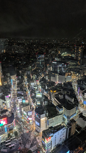 Aerial view of a vibrant cityscape at night. Colorful neon signs and busy streets with pedestrians. A bustling, lively urban atmosphere. Shibuya crossing below